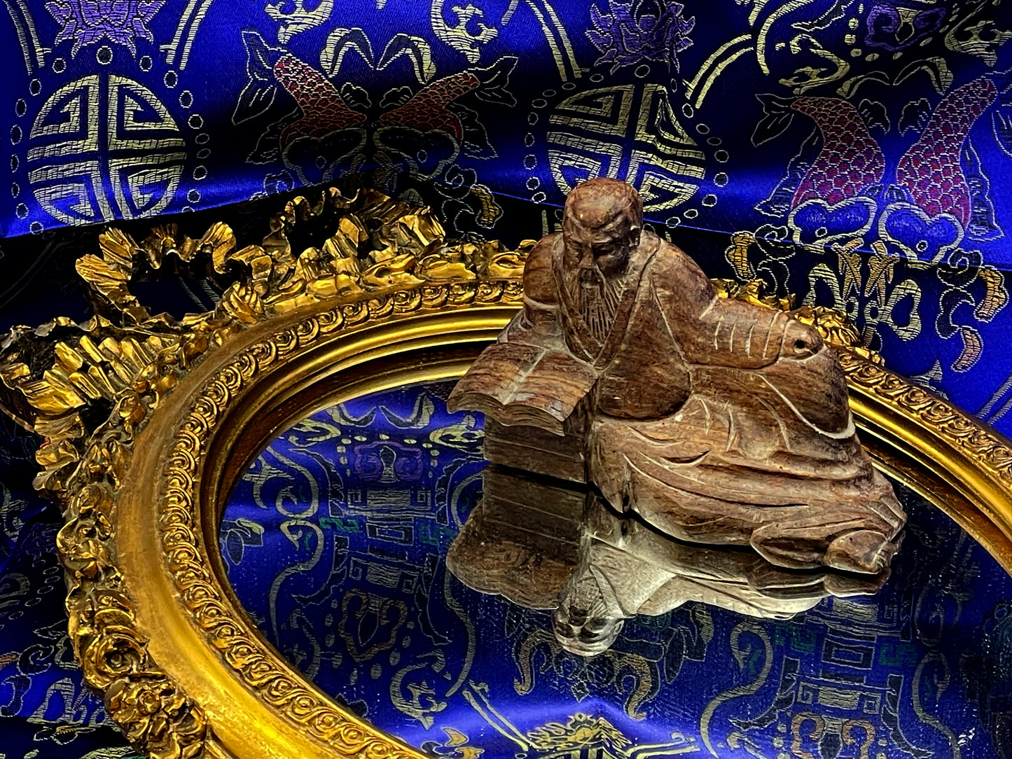 Antique Chinese Confucius hardwood sculpture beside a richly carved gilt oval mirror on blue silk