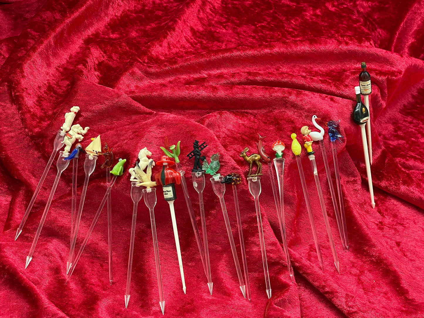 Full fanned array of vintage figural cocktail picks on red velvet, glass and celluloid toppers