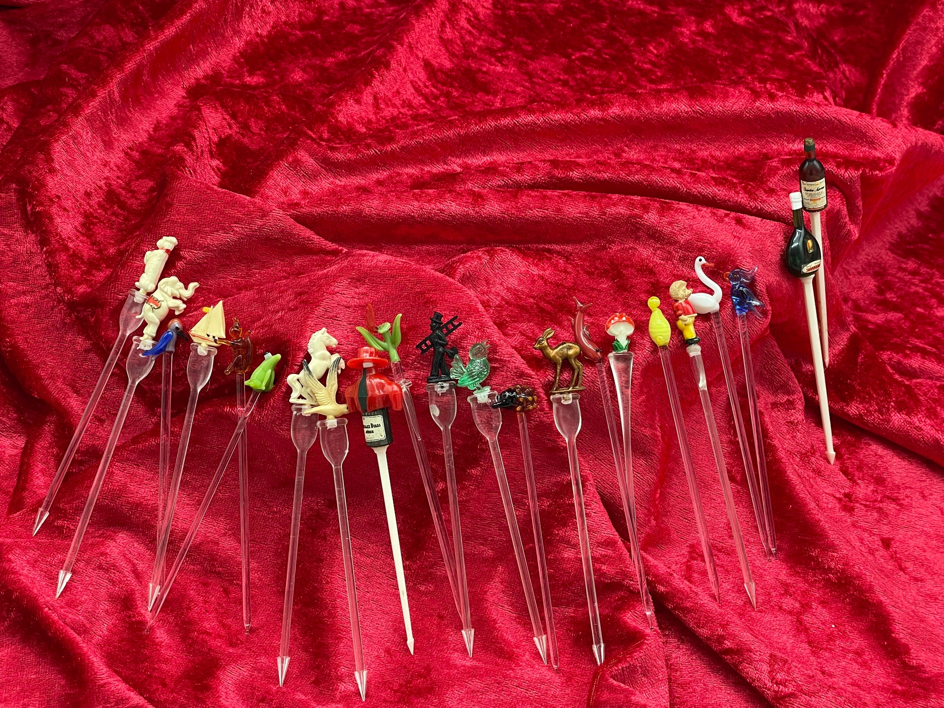 Full fanned array of vintage figural cocktail picks on red velvet, glass and celluloid toppers
