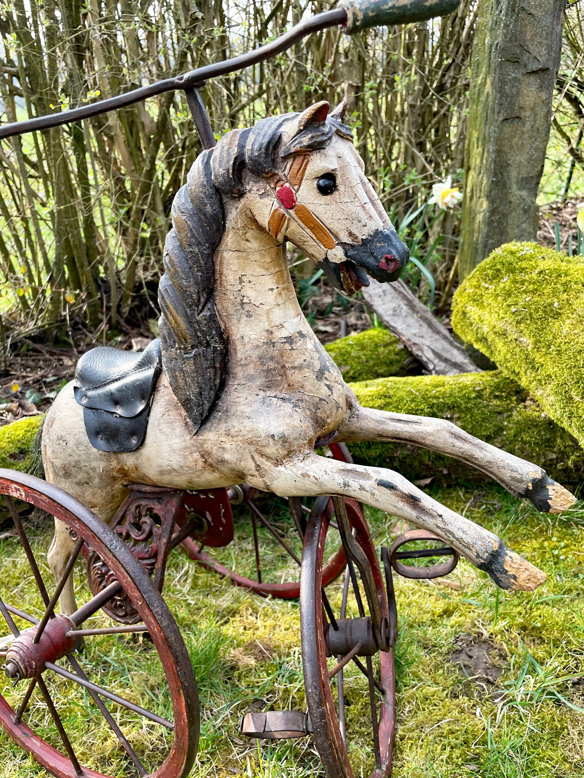 Full view of the carved wooden horse tricycle in a garden, rearing pose with spoked wheels and leather saddle