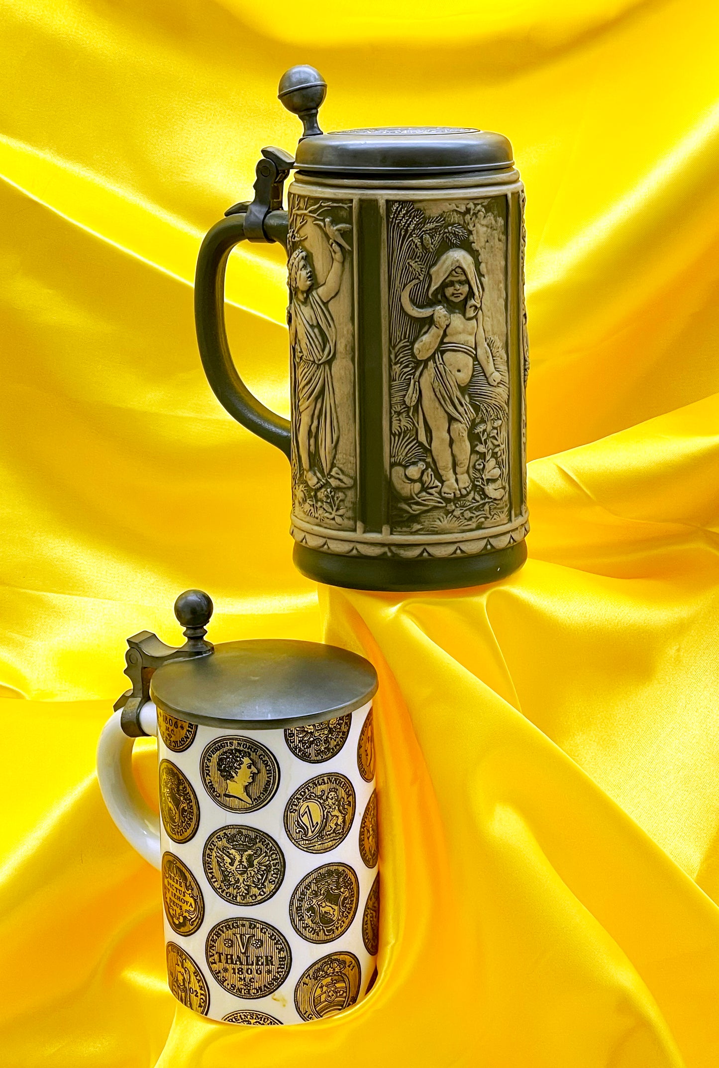 Pair of antique German beer steins with pewter lids on yellow satin, relief stein and gilt Thaler coin stein
