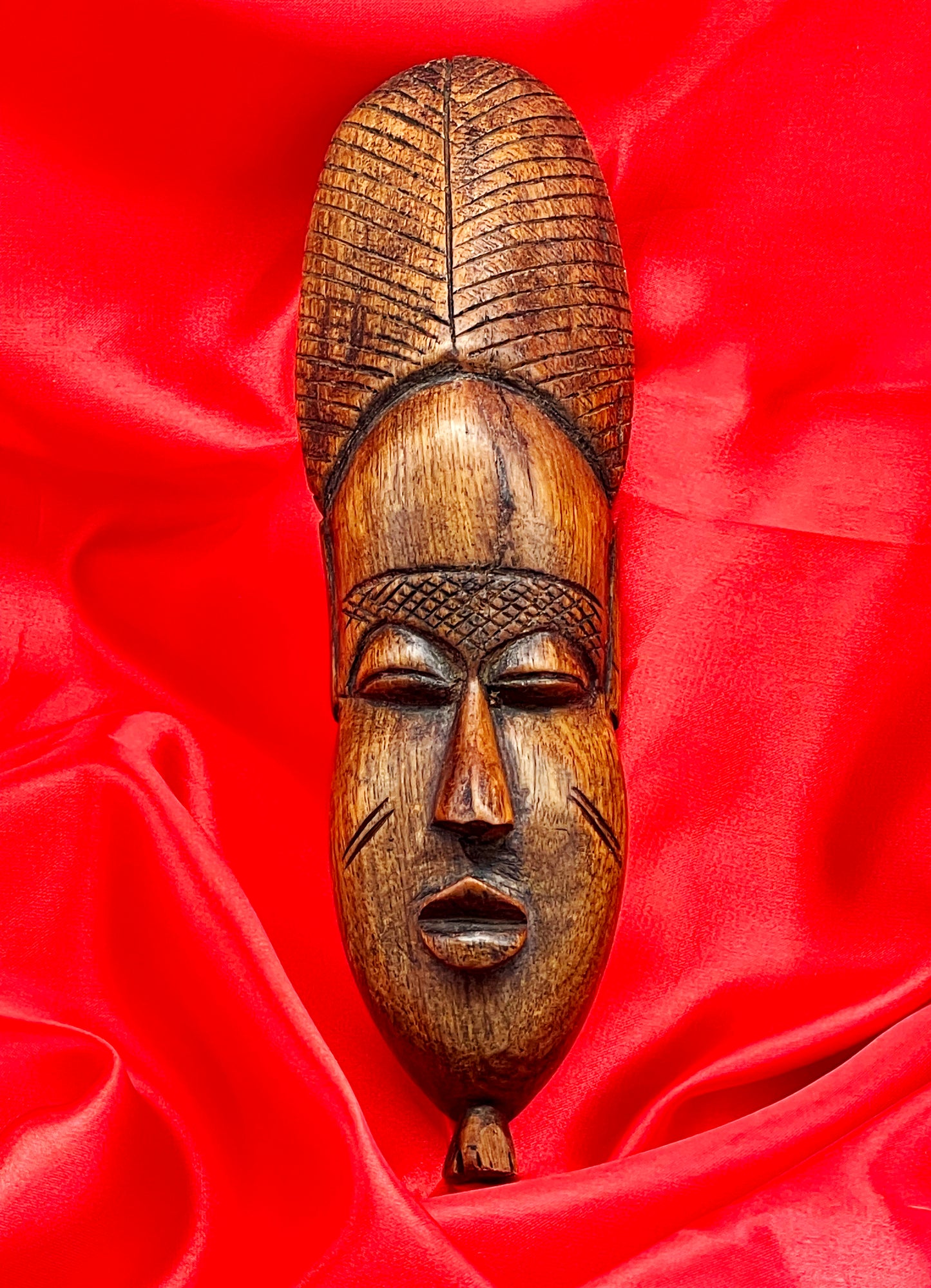 Vintage African carved hardwood mask with elongated face and ridged coiffure, front view on red ground